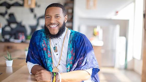 Rico Robinson stands in a café. He is wearing a blue-patterned wrap over a white button-up shirt. He is smiling.