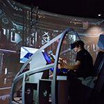 一名戴着虚拟现实头盔的学生坐在航天飞机着陆模拟器中. 他们面前是一个展示仓库的大屏幕.